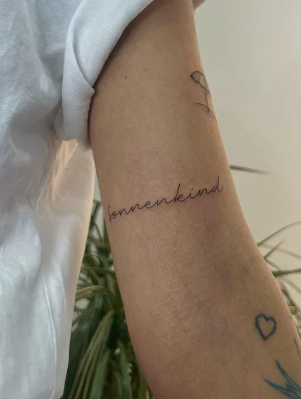 Fine Line Script 'Sonnenkind' with Heart & Flower tattoo idea for women on forearm
