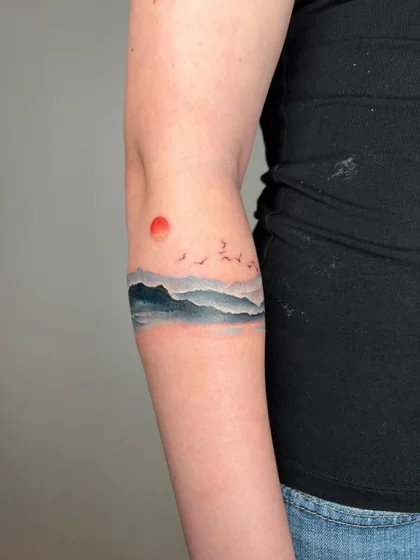 Watercolor mountain, sun, bird fine line tattoo design for women on upper arm